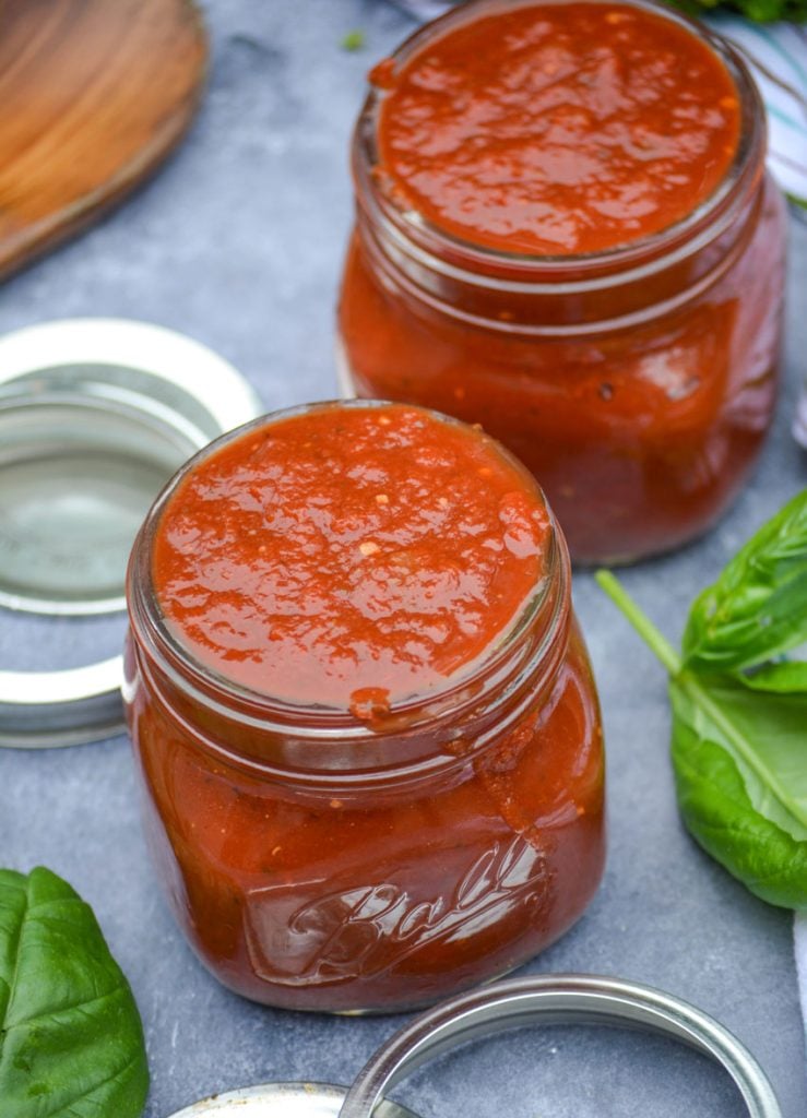 homemade marinara sauce in clear glass jars with lids and rings pictured and fresh basil leaves in the background