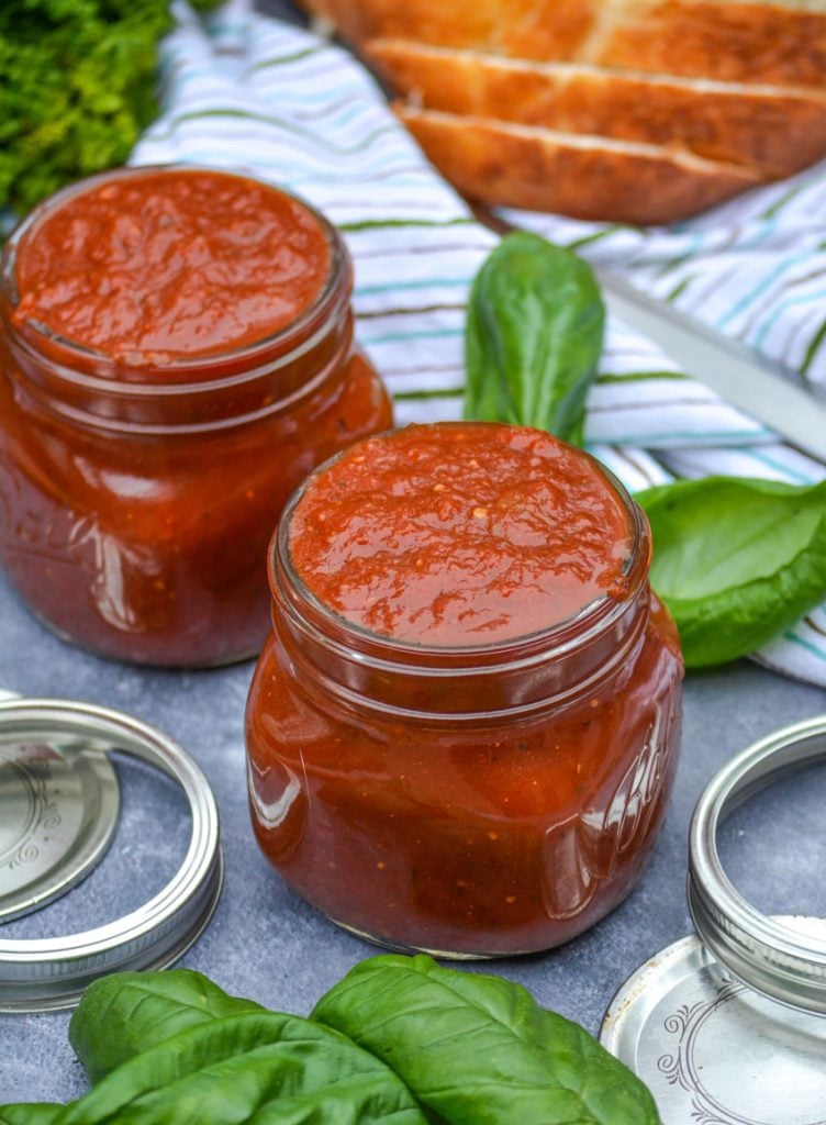 homemade marinara sauce in clear glass jars with lids and rings pictured and basil leaves and freshly sliced bread in the background