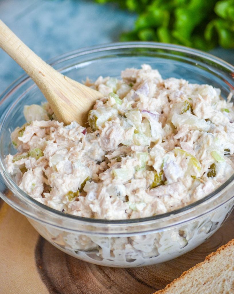creamy dill pickle tuna salad in a glass bowl on a wooden cutting board being scooped by a well worn family wooden spoon with leafy green lettuce in the background