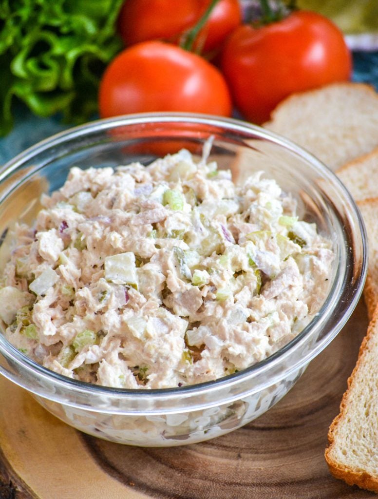 Rich & creamy, dill pickle tuna salad is in a glass bowl on a brown cutting board with freshly sliced bread green leaf lettuce and vine ripened tomatoes in the background