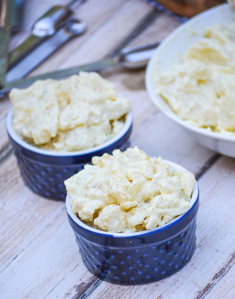 Grandma's Simple Southern Potato Salad is shown in navy blue ramekins with measuring spoons in the background on a picnic table