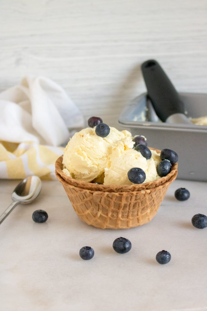 homemade no churn lemonade ice cream scoops sit in a waffle cone bowl set inside another waffle cone bowl with fresh ripe blueberries sprinkled on top