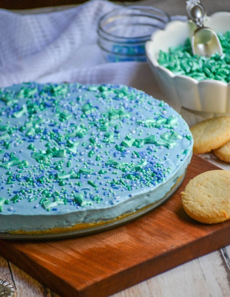 a blue hued, sprinkle topped cheesecake sitting on a dark brown cutting board with a white sail-like cloth in the back ground