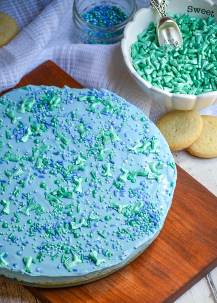 no bake mermaid cheesecake sitting on a wooden cutting board with brightly colored sprinkles in the background