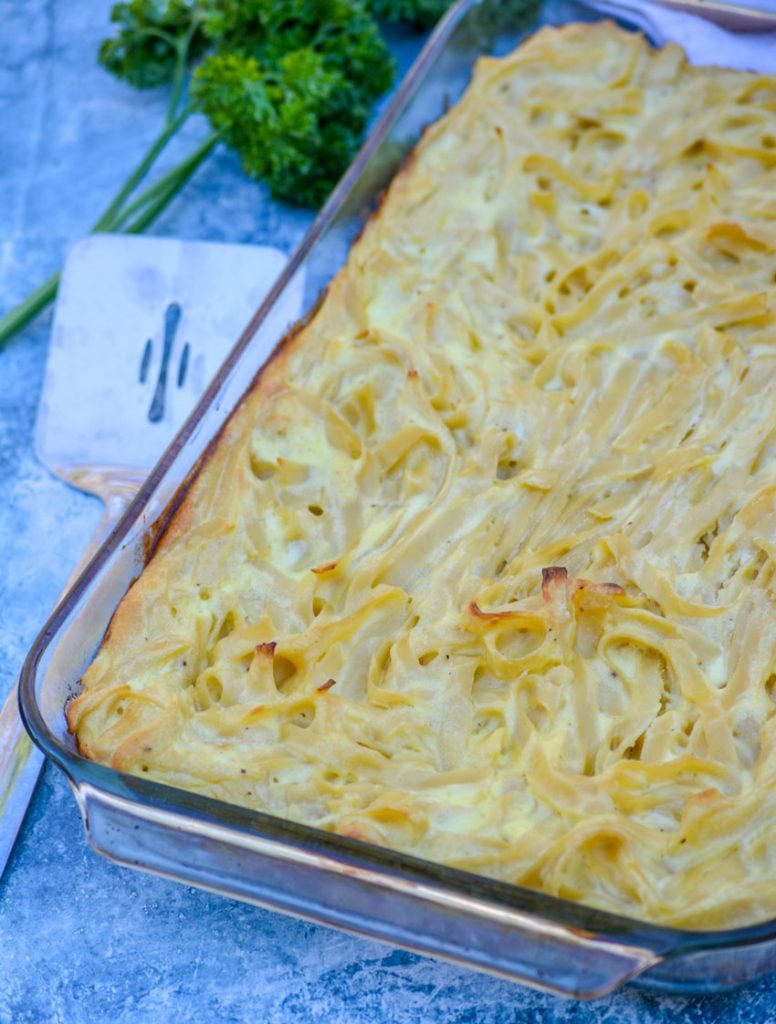 Nonna's Italian macaroni pie is shown in a glass baking fish on a light blue marbled background with a spatula ready and fresh parsley in the background