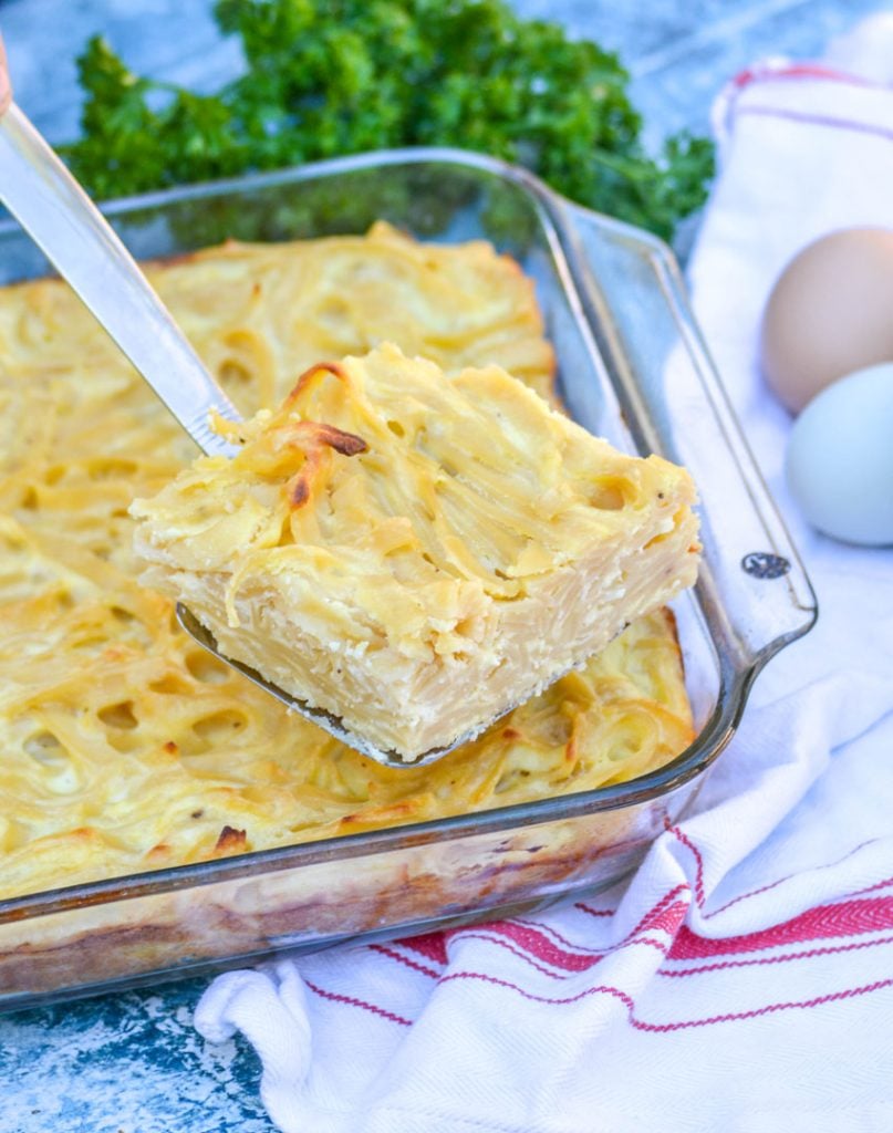 a square of this creamy Italian pasta casserole is head aloft with farm fresh eggs and bright green parsley in the background on top of a red and white cloth napkin