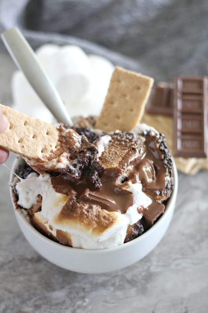 a crisp graham cracker is being dipped into a bowl of s'mores cobbler showing its decadent chocolate cake layer with toasted marshmallows and melted chocolate candy bars