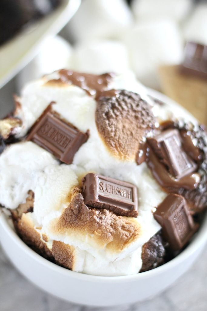 toasted marshmallows & melty mini chocolate bars top a smores cobbler in a white bowl with a casserole dish full in the background
