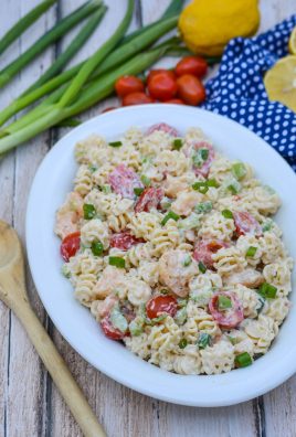 a creamy seafood style pasta salad in an oval shaped white bowl, surrounded bright fresh veggies on a picnic table with a weathered wooden spoon for serving
