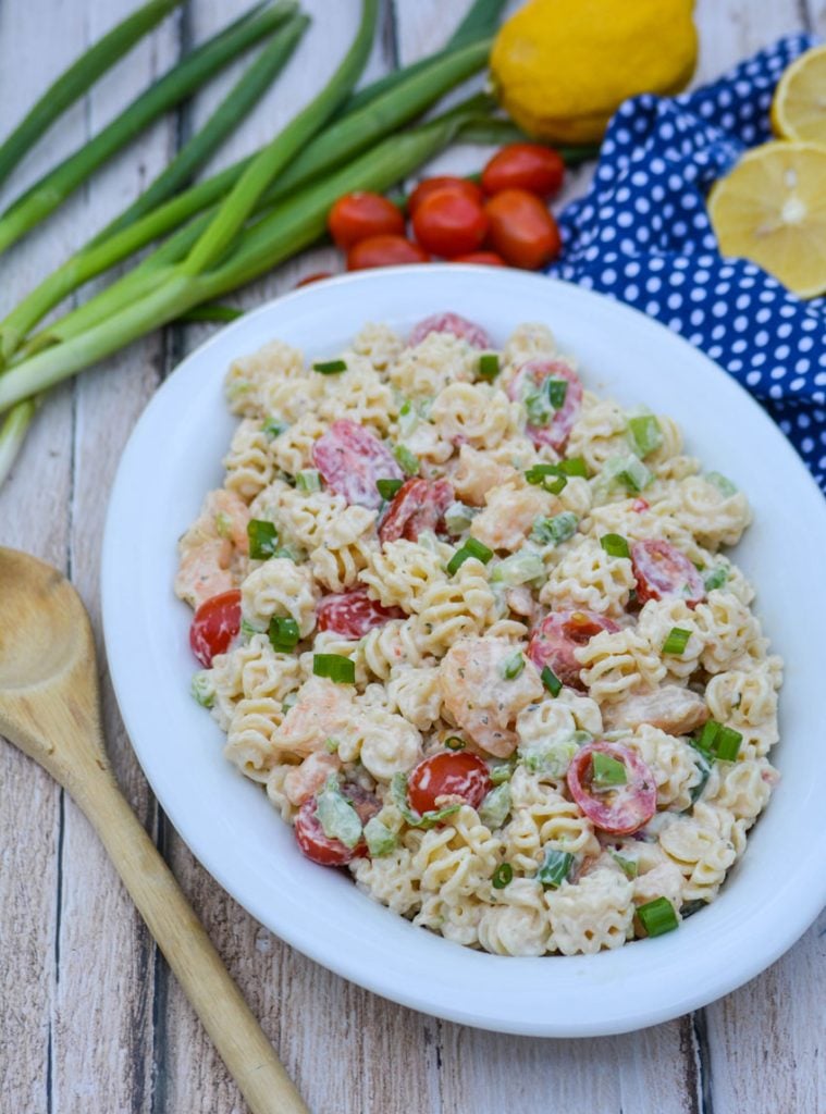 a creamy seafood style pasta salad in an oval shaped white bowl, surrounded bright fresh veggies on a picnic table with a weathered wooden spoon for serving