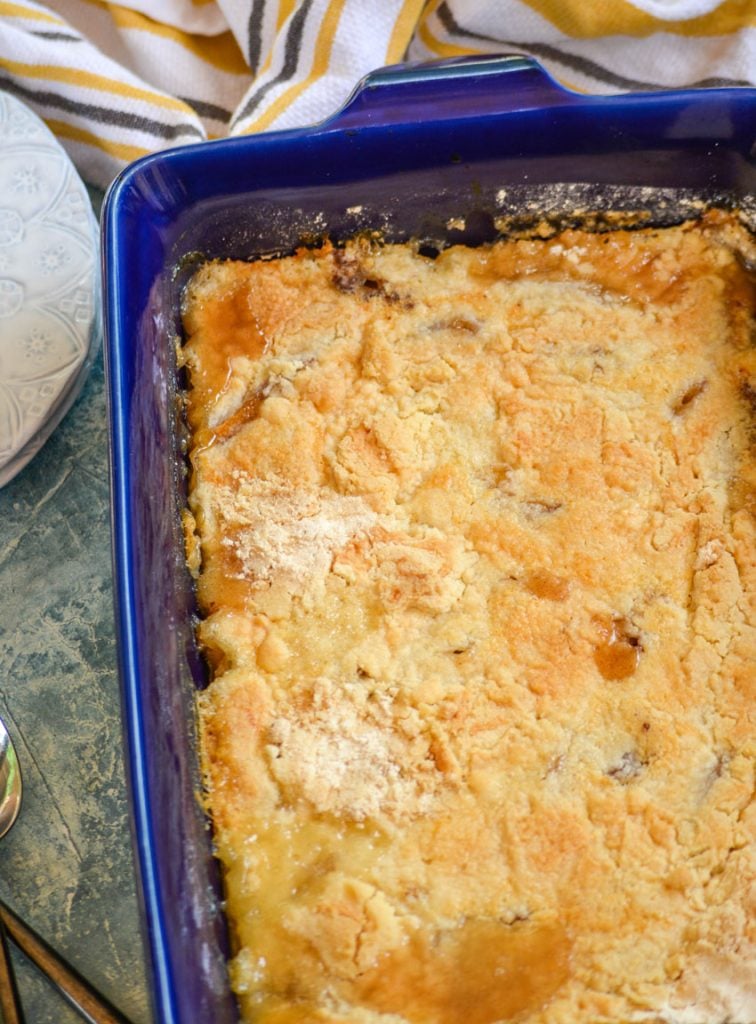 The peach cobbler dump cake recipe is baked in a royal blue rectangular dish on a faded teal marbled counter top with white plates in the background