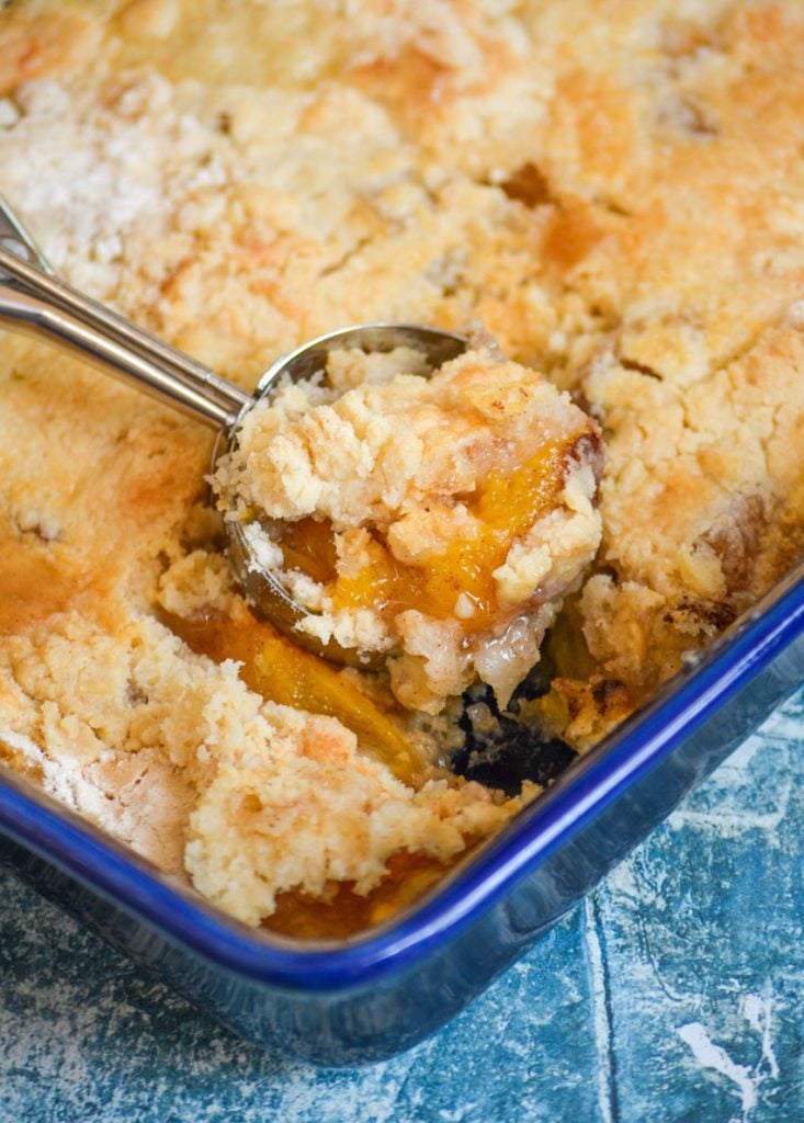 a metal ice cream scooper is full of peach cobbler dump cake, the peaches are glistening and the cobbler layer looks crumbly