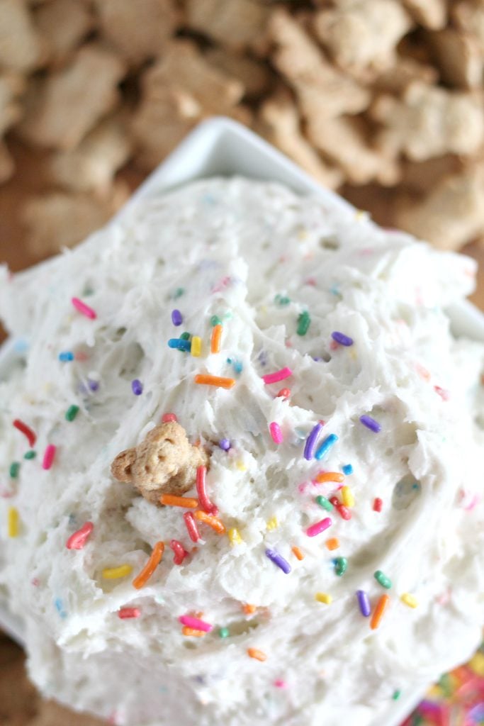 a fluffy white cake batter dip is flecked with rainbow sprinkles and piled high in a white square bowl with teddy grahams spread out in the background