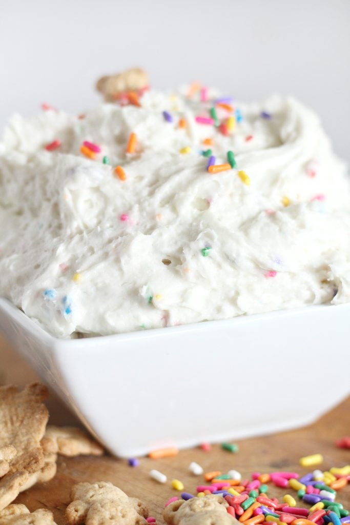fluffy dunkaroo dip is shown in a bright white square bowl sprinkled with rainbow colored sprinkles with a cute teddy graham sticking out the top at an angle