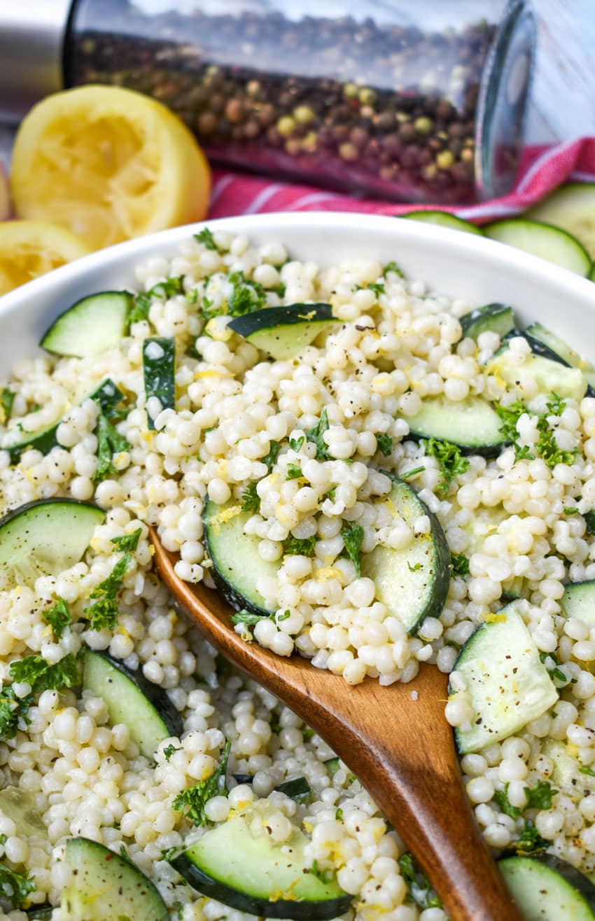 a wooden spoon stuck in a white bowl filled with cucumber couscous salad