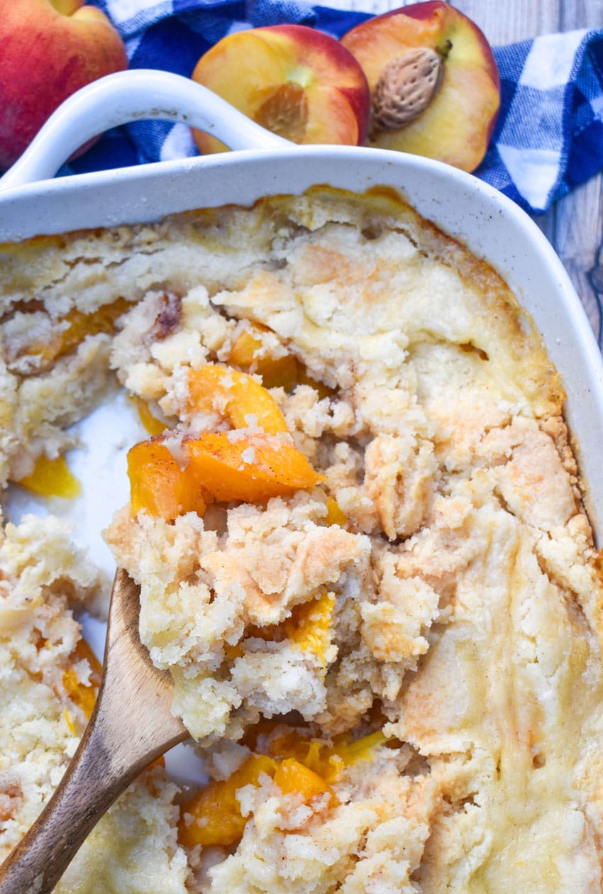 a wooden spoon scooping peach cobbler dump cake out of a white baking dish
