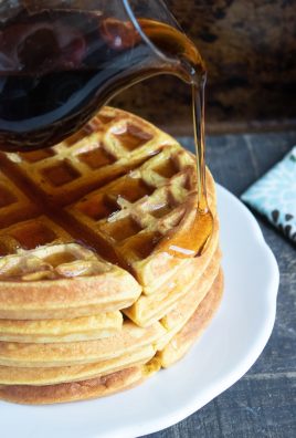 a fluffy stack of lightly spiced pumpkin flavored waffles on a white plate is shown with rich syrup being slowly poured on top and dripping down the edge