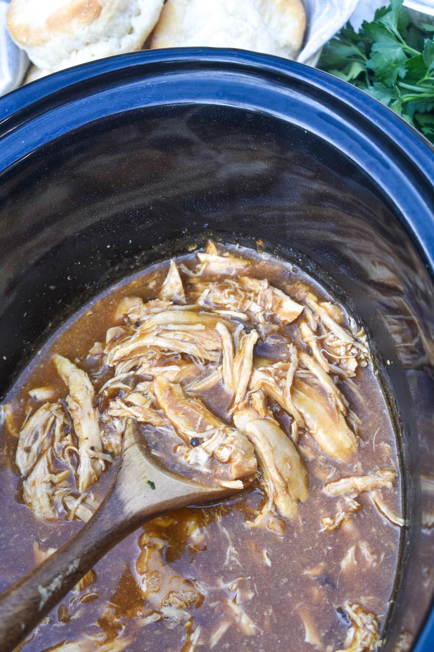 a wooden spoon scooping shredded chicken and brown gravy out of a slow cooker