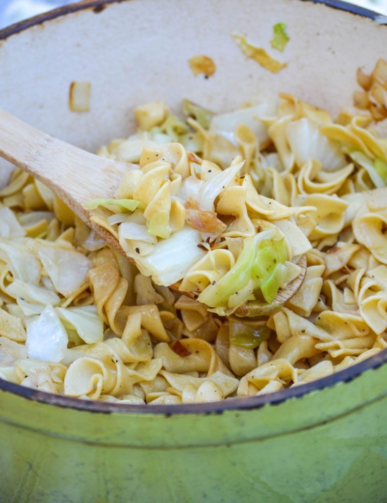 a wooden spoon scooping Slovak Haluski Recipe (Polish fried cabbage & noodles) out of a cast iron Dutch oven pot