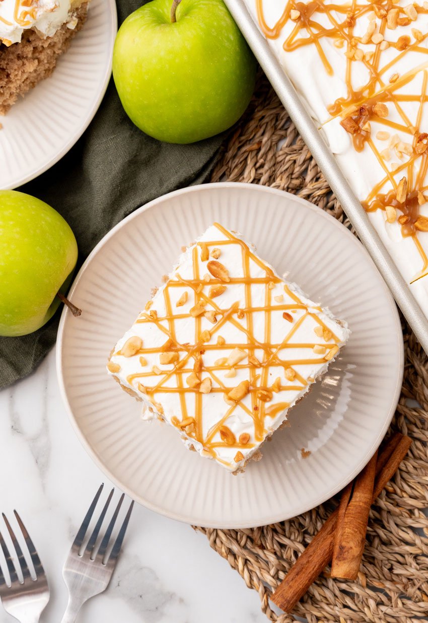 A SLICE OF AN APPLE SPICE POKE CAKE ON A SMALL WHITE PLATE WITH A GREEN APPLE IN THE BACKGROUND