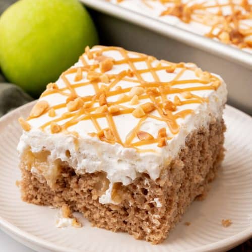 A SLICE OF AN APPLE SPICE POKE CAKE ON A SMALL WHITE PLATE WITH A GREEN APPLE IN THE BACKGROUND