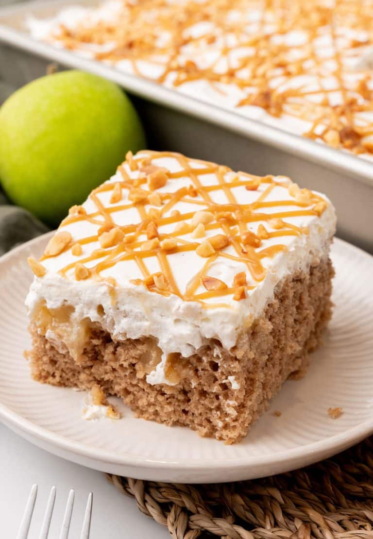 A SLICE OF AN APPLE SPICE POKE CAKE ON A SMALL WHITE PLATE WITH A GREEN APPLE IN THE BACKGROUND