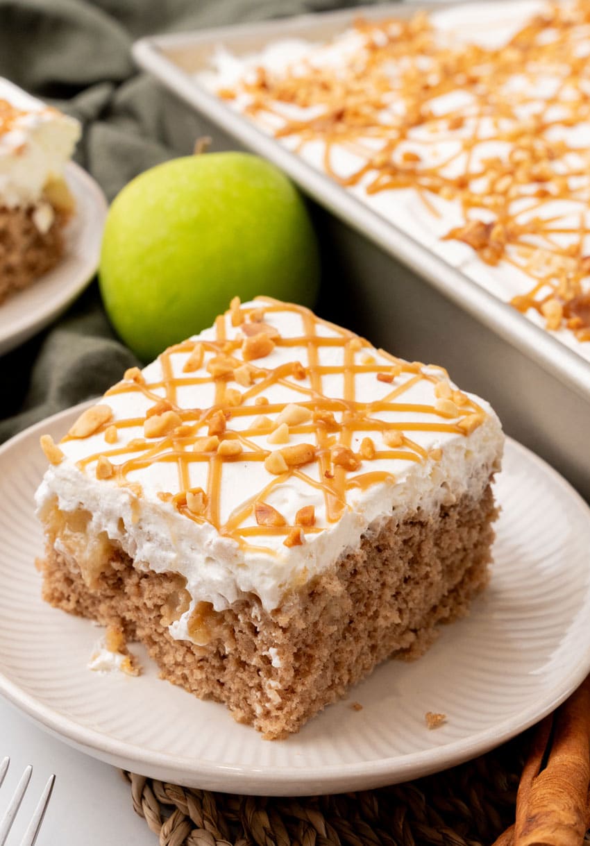 A SLICE OF AN APPLE SPICE POKE CAKE ON A SMALL WHITE PLATE WITH A GREEN APPLE IN THE BACKGROUND