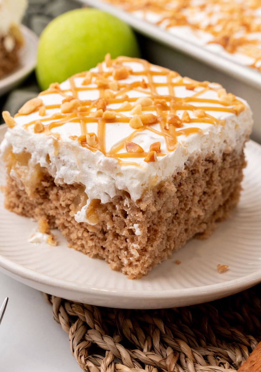 A SLICE OF AN APPLE SPICE POKE CAKE ON A SMALL WHITE PLATE WITH A GREEN APPLE IN THE BACKGROUND