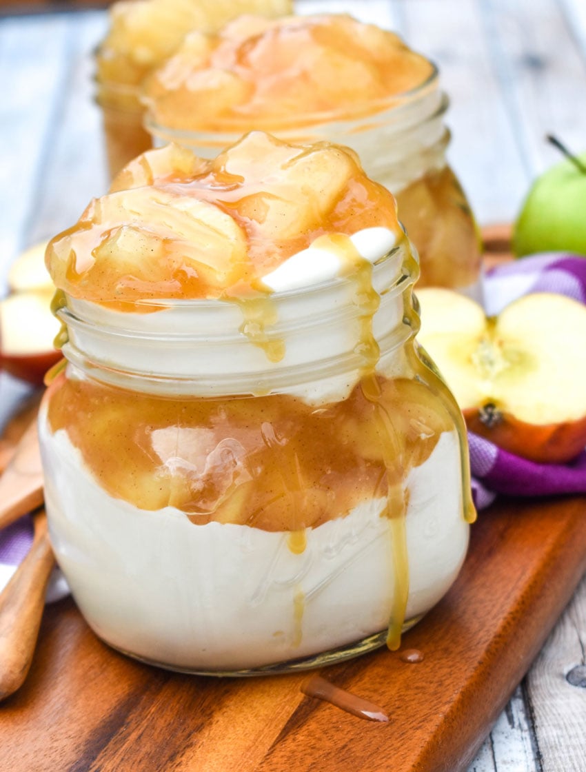 two mason jar apple cheesecake parfaits drizzled with caramel syrup on a wooden cutting board