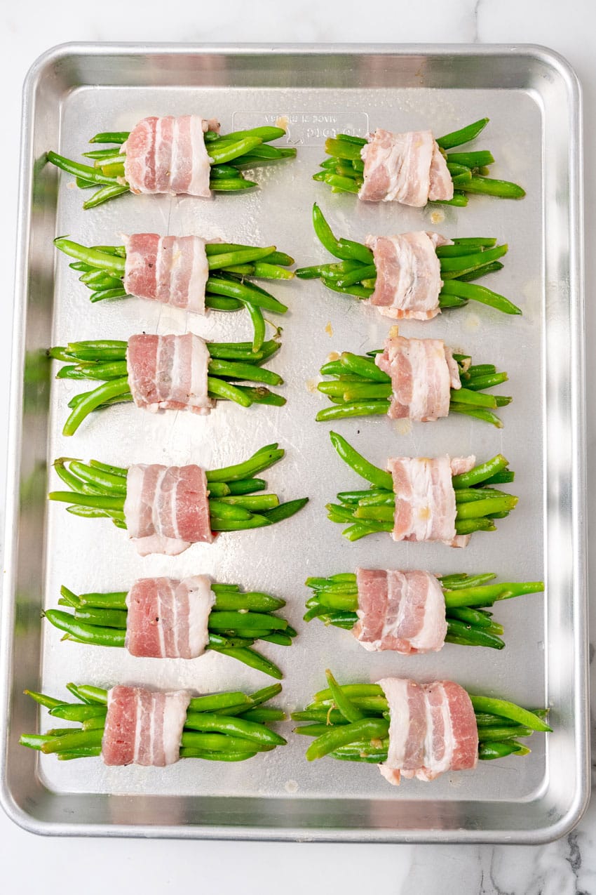 bacon wrapped green bean bundles arranged in two rows on a metal baking pan