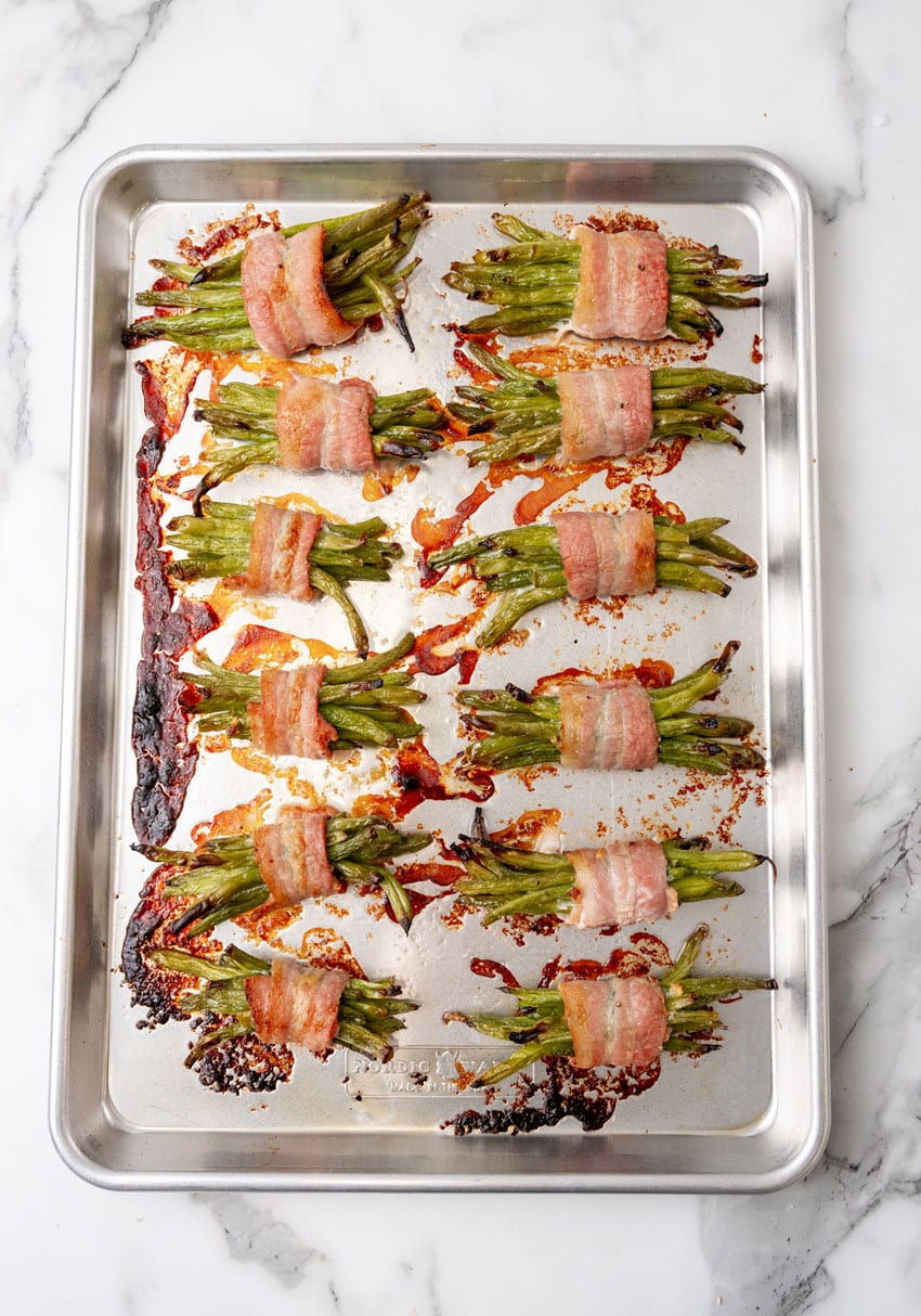 roasted bacon wrapped green bean bundles arranged in two rows on a metal baking pan
