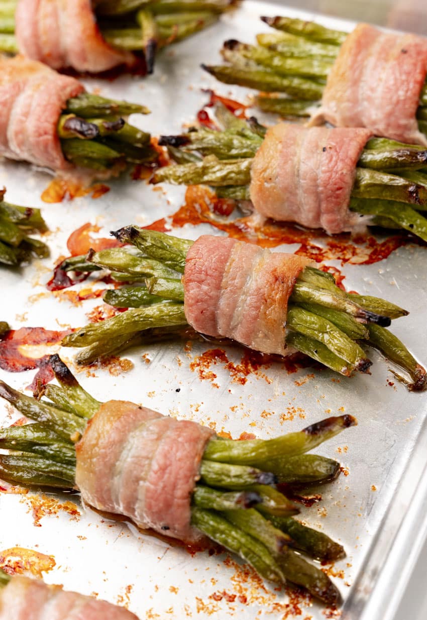roasted bacon wrapped green bean bundles arranged in two rows on a metal baking pan