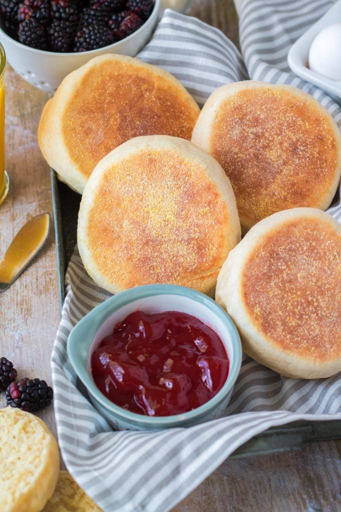 homemade english muffins in a cloth lined metal tray with a bowl of cherry jam