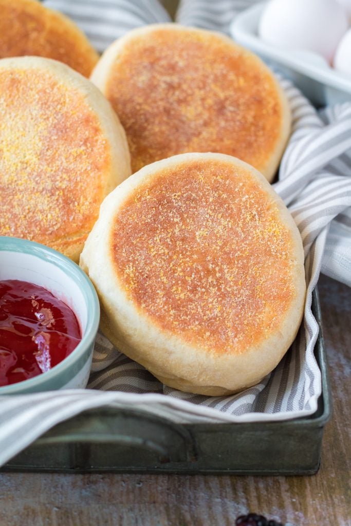 homemade english muffins in a cloth lined metal tray with a bowl of cherry jam