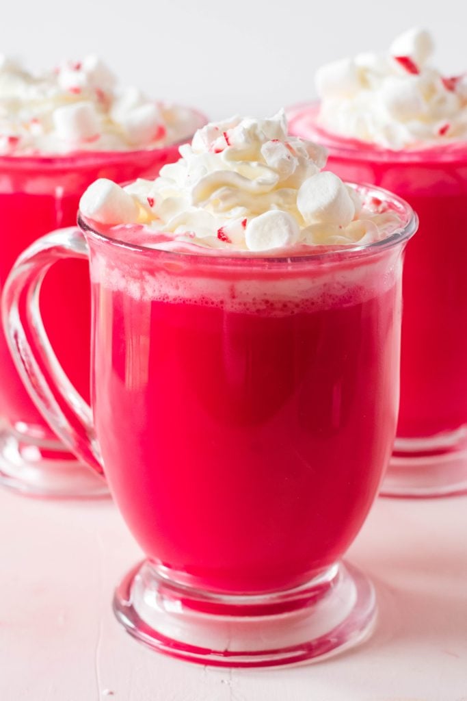 rich red velvet hot chocolate is pictured in a clear glass mug and topped with whipped cream and sprinkled with mini marshmallows and crushed candy cane bits