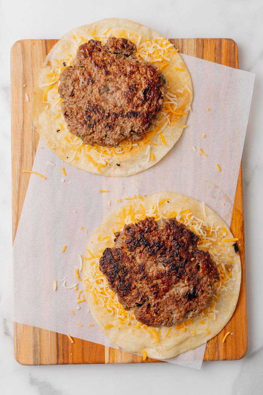 crispy flour tortillas topped with shredded cheese and burger patties on a parchment paper lined wooden cutting board