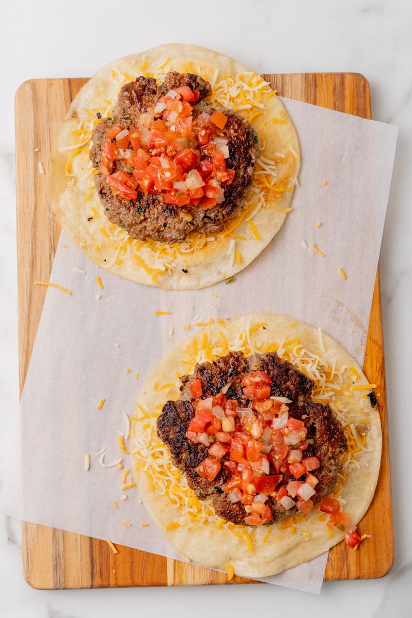 crispy flour tortillas topped with shredded cheese, burger patties, and pico de gallo on a parchment paper lined wooden cutting board