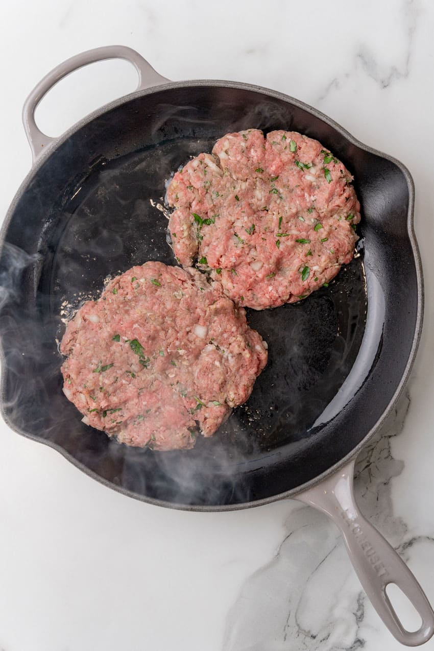two burger patties cooking in a large black skillet