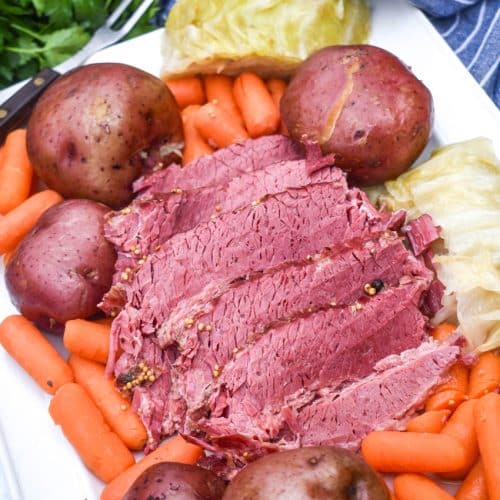 slices of cooked corned beef brisket on a white platter surrounded by potatoes cabbage and carrots