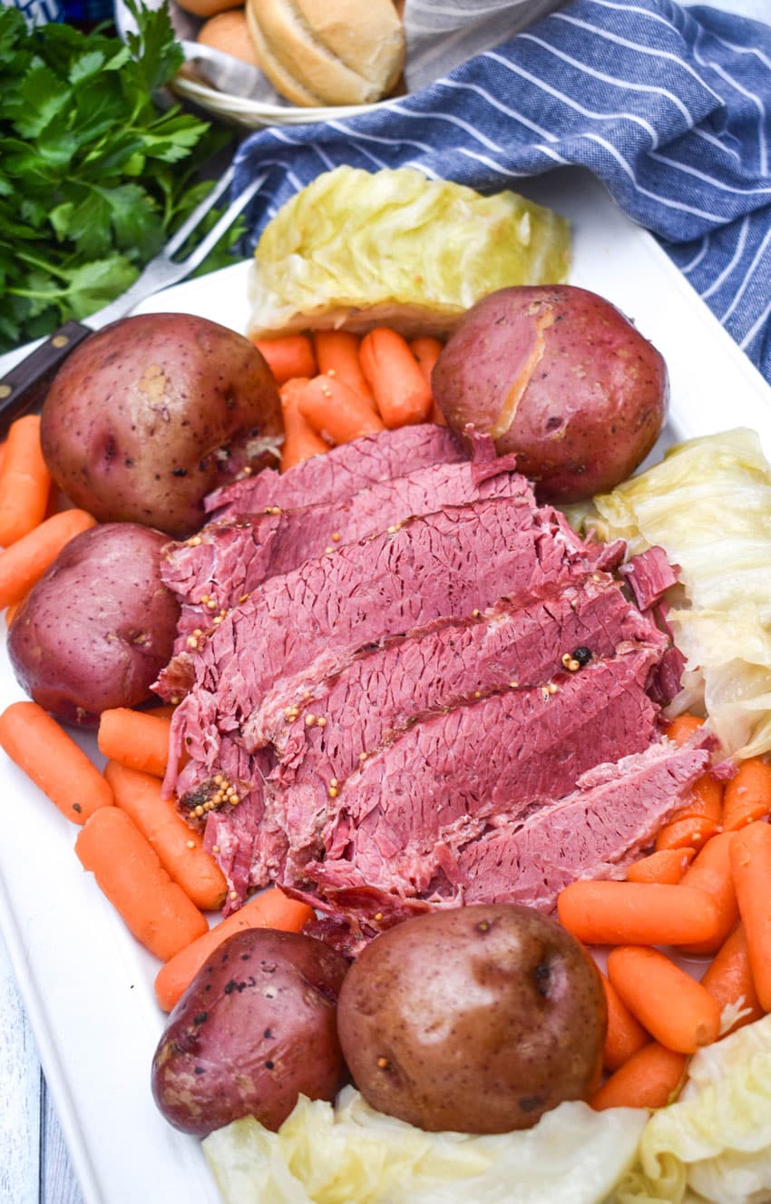 slices of cooked corned beef brisket on a white platter surrounded by potatoes cabbage and carrots