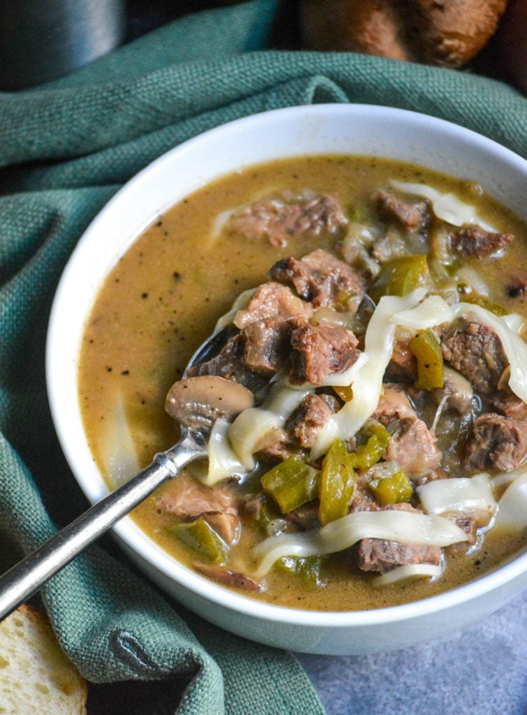 philly cheesesteak stew served in a white soup bowl with a metal spoon shown scooping some up