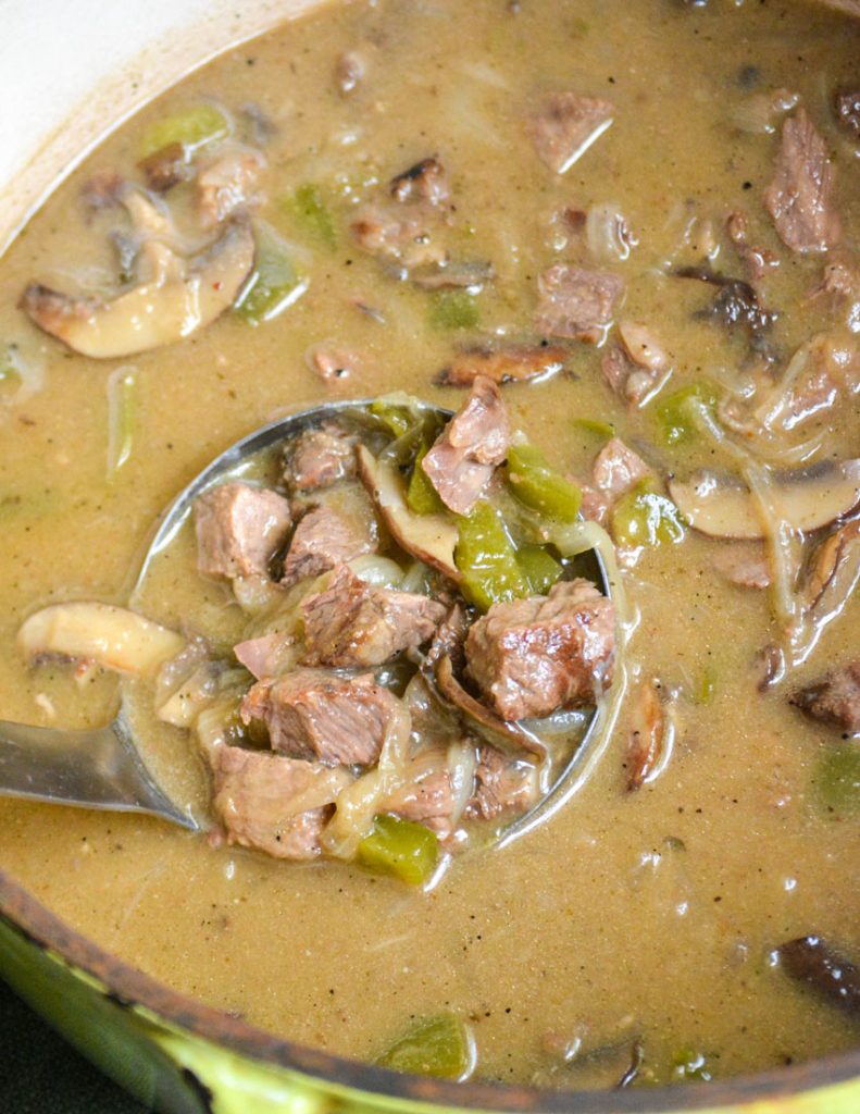 a silver ladle shown with a scoop of cheesesteak soup in a cast iron Dutch oven