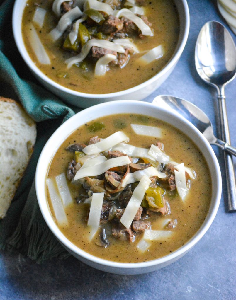 philly cheesesteak stew shown in white soup bowls topped with strips of provolone cheese with a spoon and sliced bread on the side