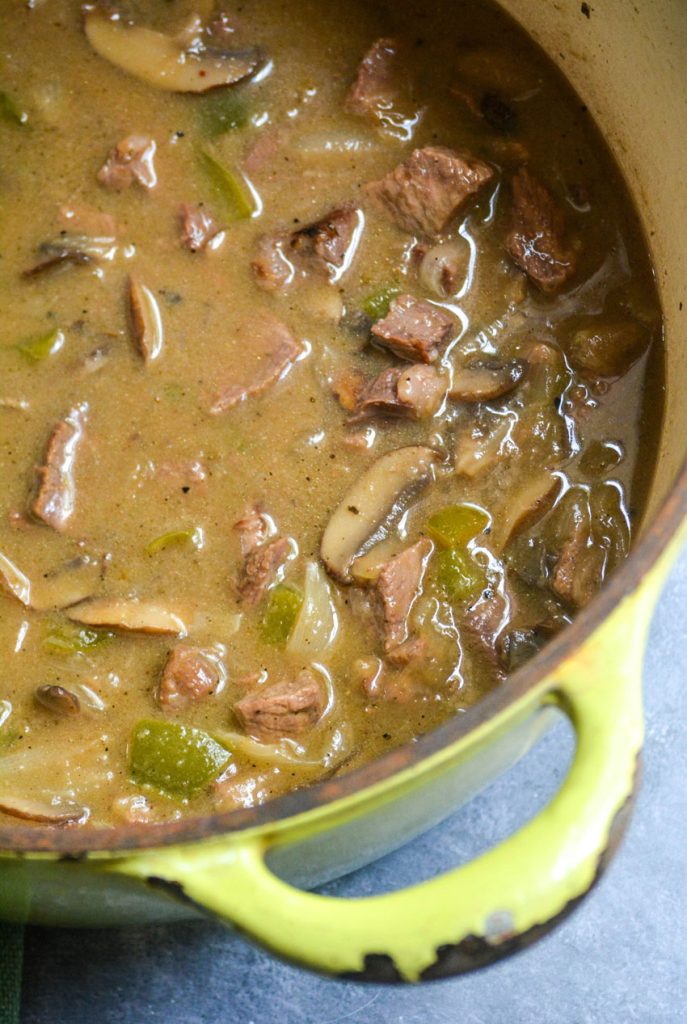 mushrooms, green bell peppers, onions and chunks of cooked steak are shown floating in a rich brown broth inside a green dutch oven