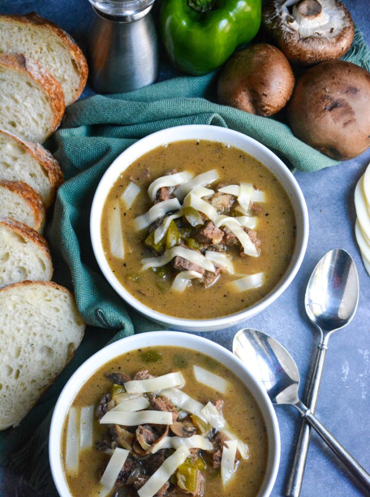 philly cheesesteak stew shown in white soup bowls topped with strips of provolone cheese with a spoon and sliced bread on the side