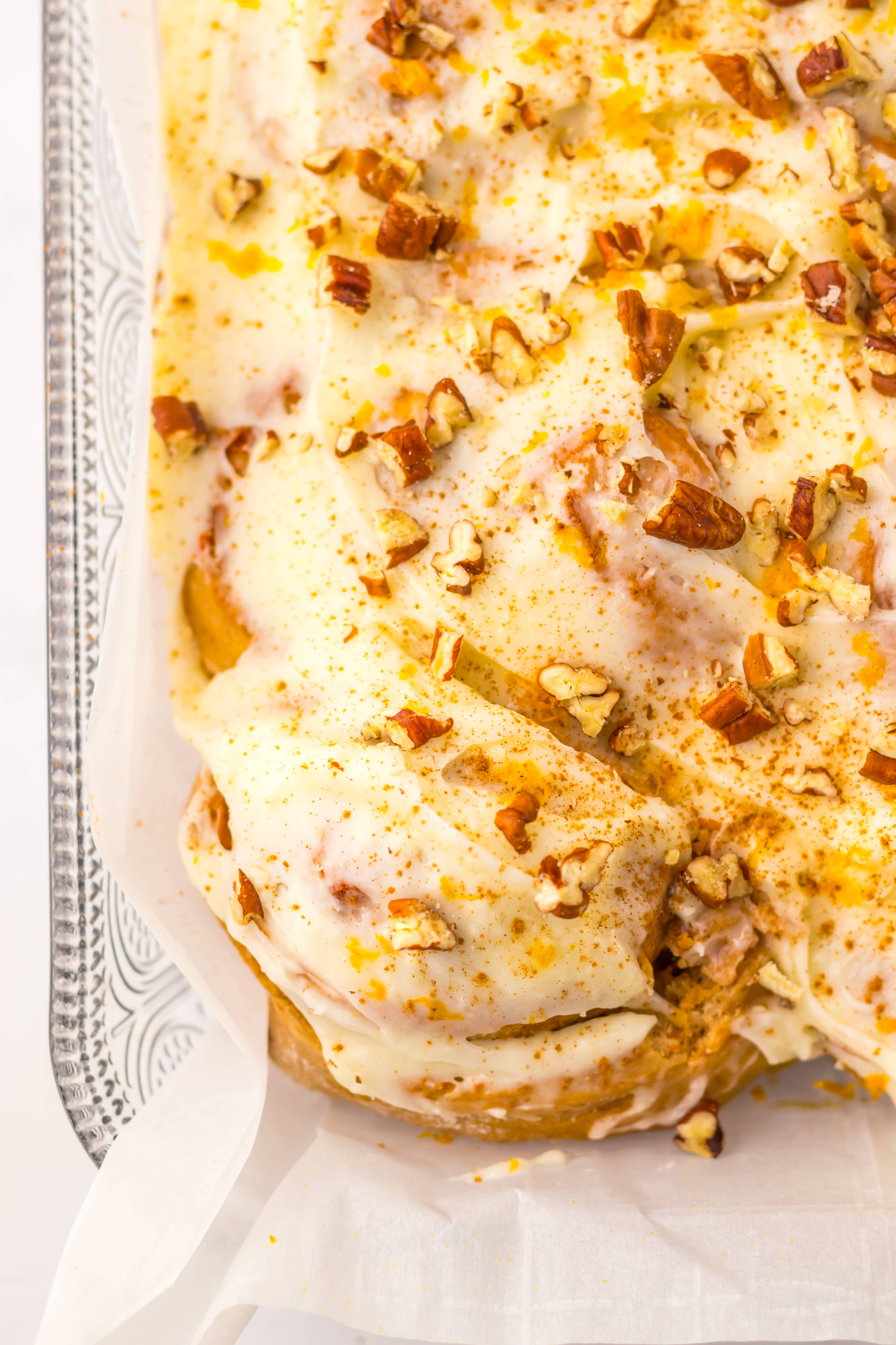 a close up of frosted carrot cake cinnamon rolls in a parchment paper lined glass baking dish
