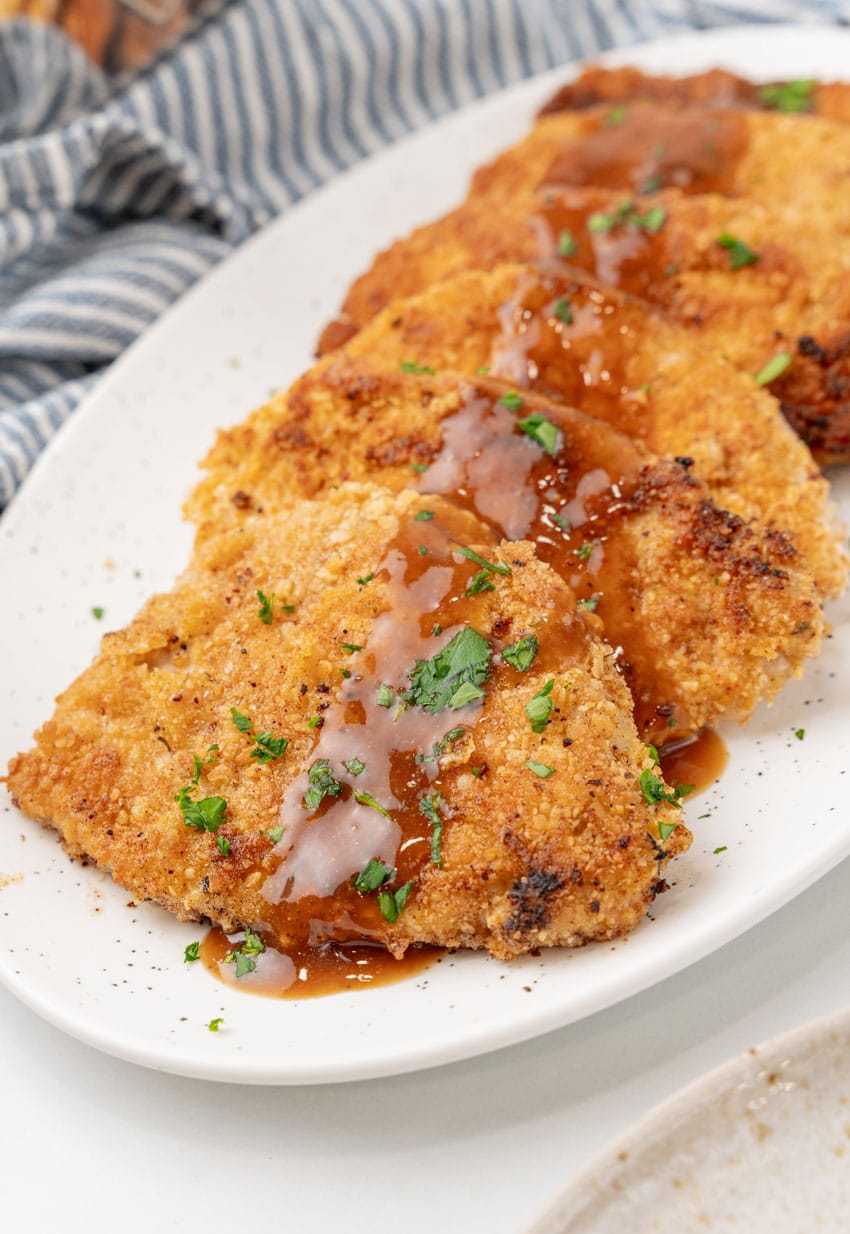 pieces og homemade pork schnitzel topped with brown gravy arranged in a row on a white serving platter