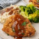 pork schnitzel topped with brown gravy on a white plate with steamed broccoli and mashed potatoes on the side