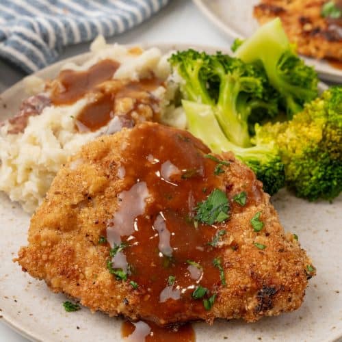 pork schnitzel topped with brown gravy on a white plate with steamed broccoli and mashed potatoes on the side