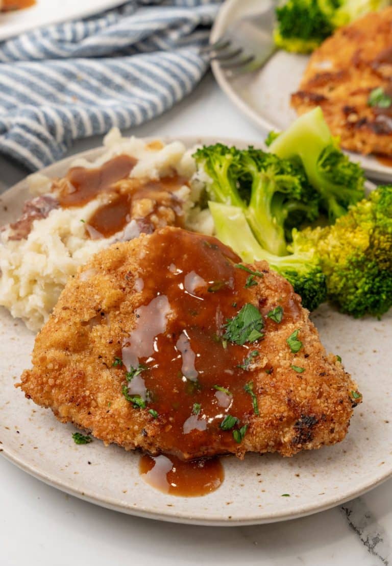 pork schnitzel topped with brown gravy on a white plate with steamed broccoli and mashed potatoes on the side